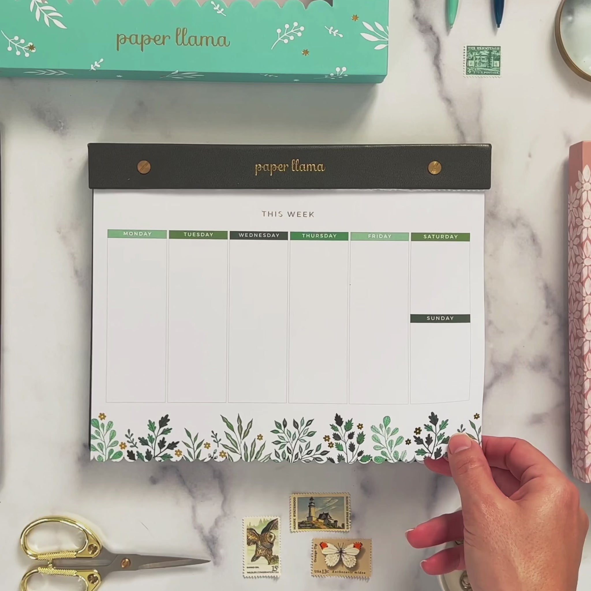 A Video with a Desktop Notepad with colorful watercolor leaves on the bottom and scalloped edge on the bottom. The middle contains a weekly planner with room to out each day. The background is a deskscape made of marble. A hands comes on the screen and lifts the first sheet of paper so you can see the foil shimmering. Then it rips the top sheet off the notepad