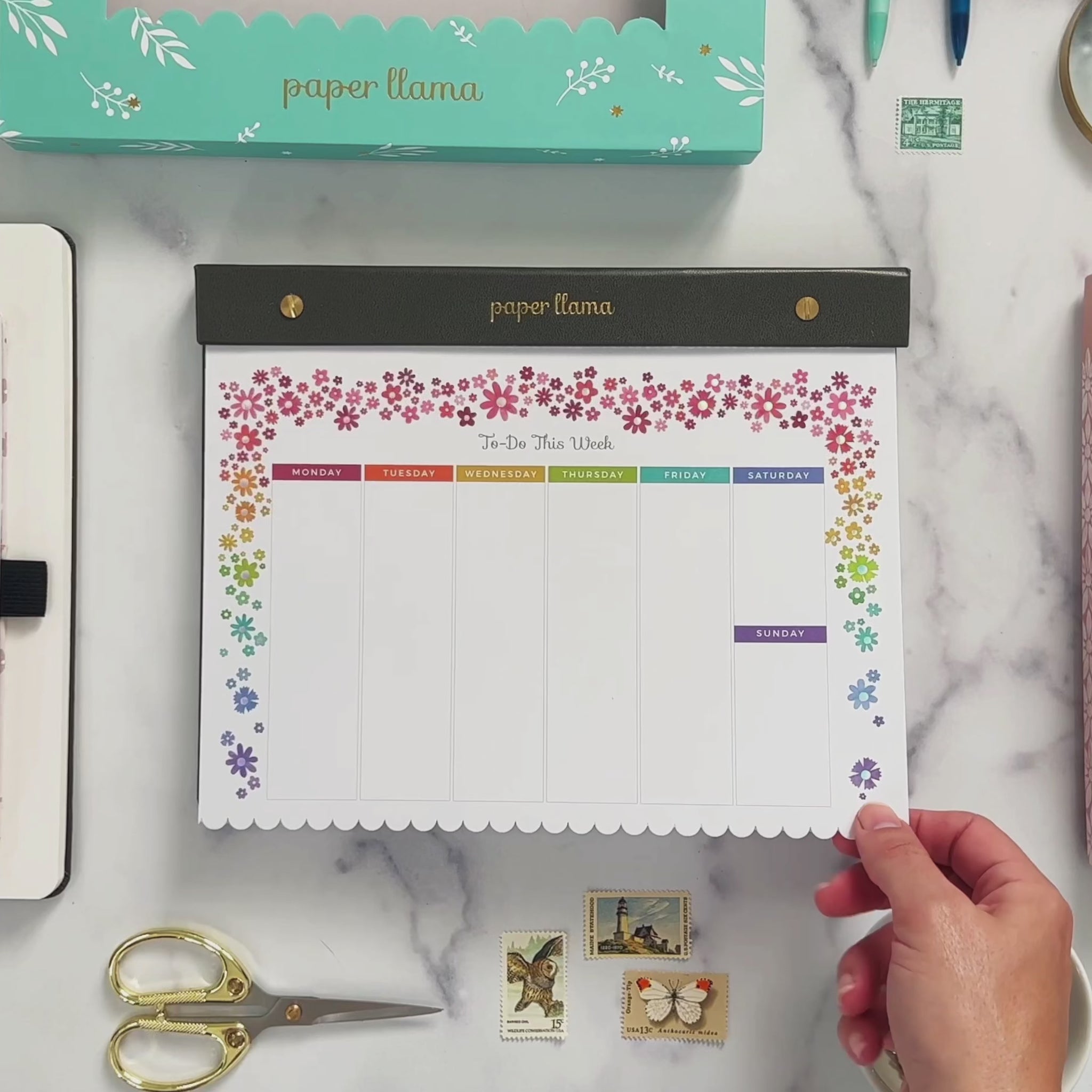 A Video with a Desktop Notepad with colorful flowers on the top and scalloped edge on the bottom. The middle contains a weekly planner with room to out each day. The background is a deskscape made of marble. A hands comes on the screen and lifts the first sheet of paper so you can see the foil shimmering. Then it rips the top sheet off the notepad