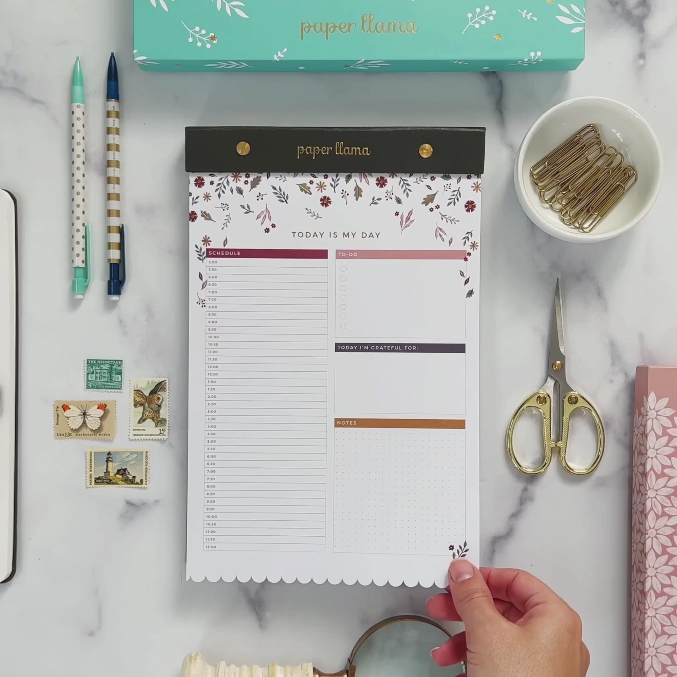 A Video with a Desktop Notepad with colorful leaves falling from the top on the top and scalloped edge on the bottom. The middle contains a daily planner with room to plan hour by hour, organize your to-do's, add gratitude, and take notes. Then the background changes to be a deskscape made of marble. A hands comes on teh screen and lifts the first sheet of paper so you can see the foil shimmering. Then it rips the top sheet off the notepad