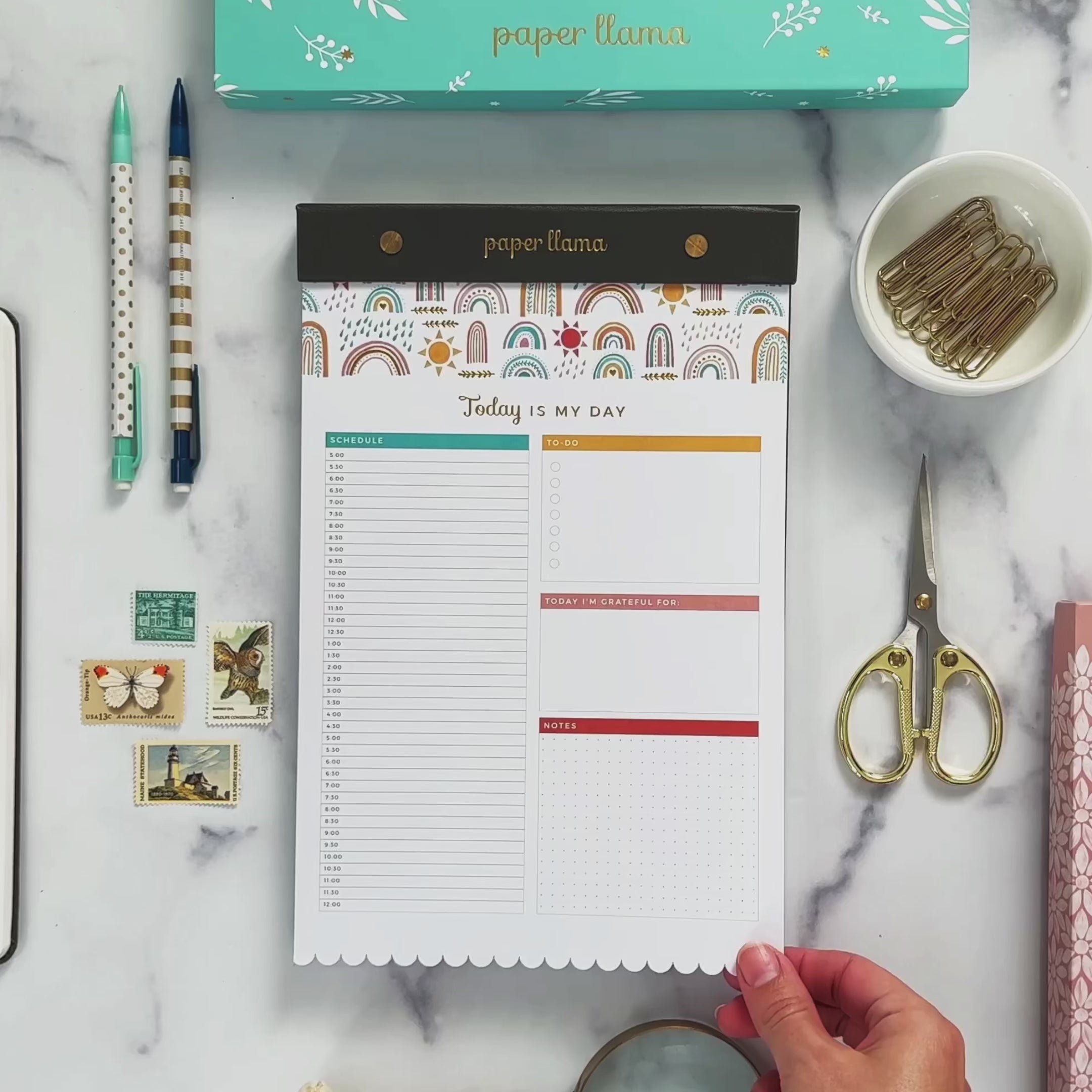 A Video with a Desktop Notepad with colorful rainbows on the top and scalloped edge on the bottom. The middle contains a daily planner with room to plan hour by hour, organize your to-do's, add gratitude, and take notes. Then the background changes to be a deskscape made of marble. A hands comes on teh screen and lifts the first sheet of paper so you can see the foil shimmering. Then it rips the top sheet off the notepad