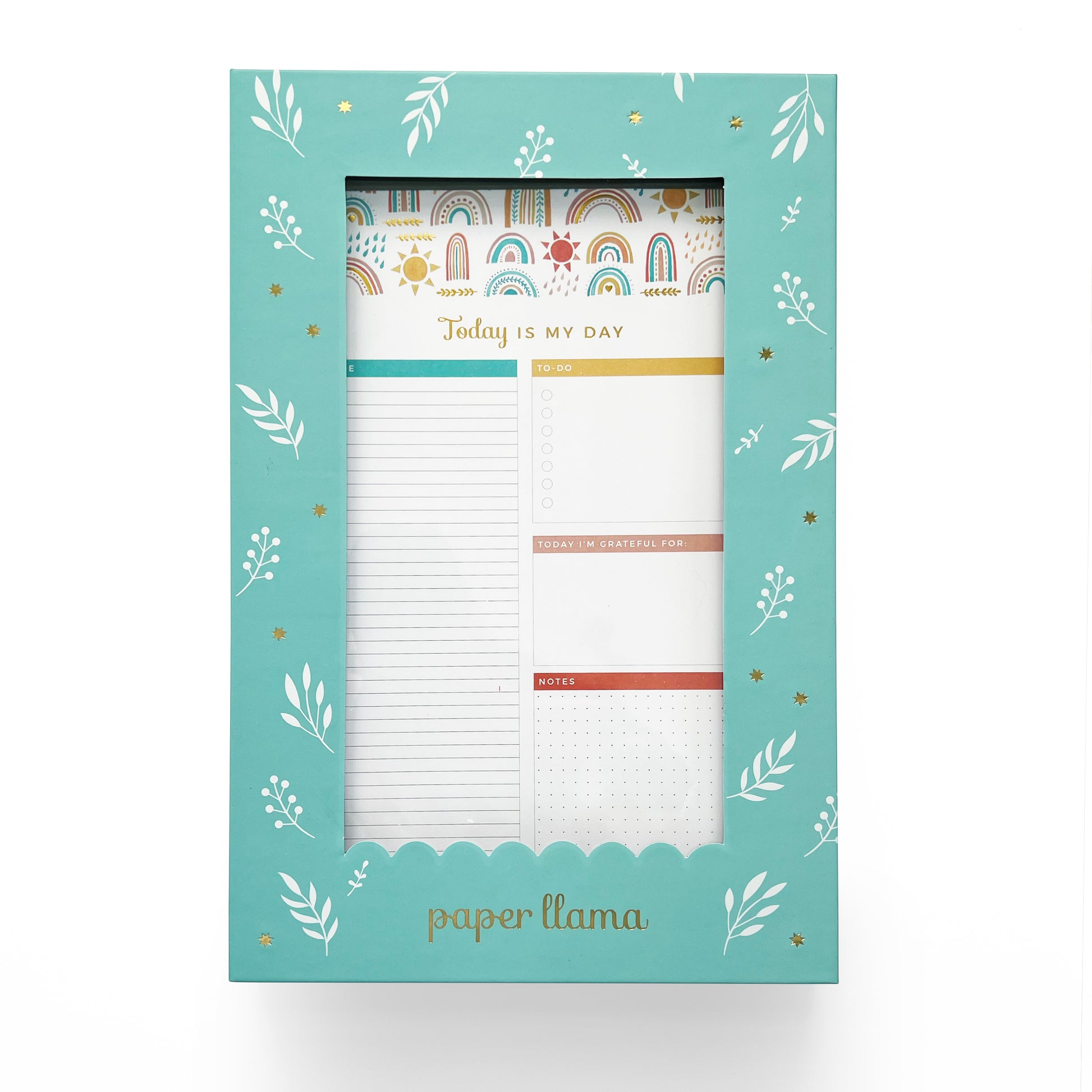 A teal blue box with white florals top. The box contains A Desktop Notepad with colorful rainbows on the top and scalloped edge on the bottom. The middle contains a daily planner with room to plan hour by hour, organize your to-do's, add gratitude, and take notes